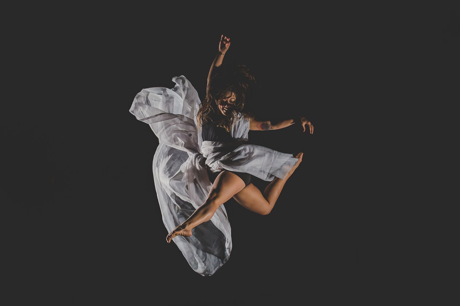 Contemporary dancer leaps through air wearing billowing white dress against dark backdrop.