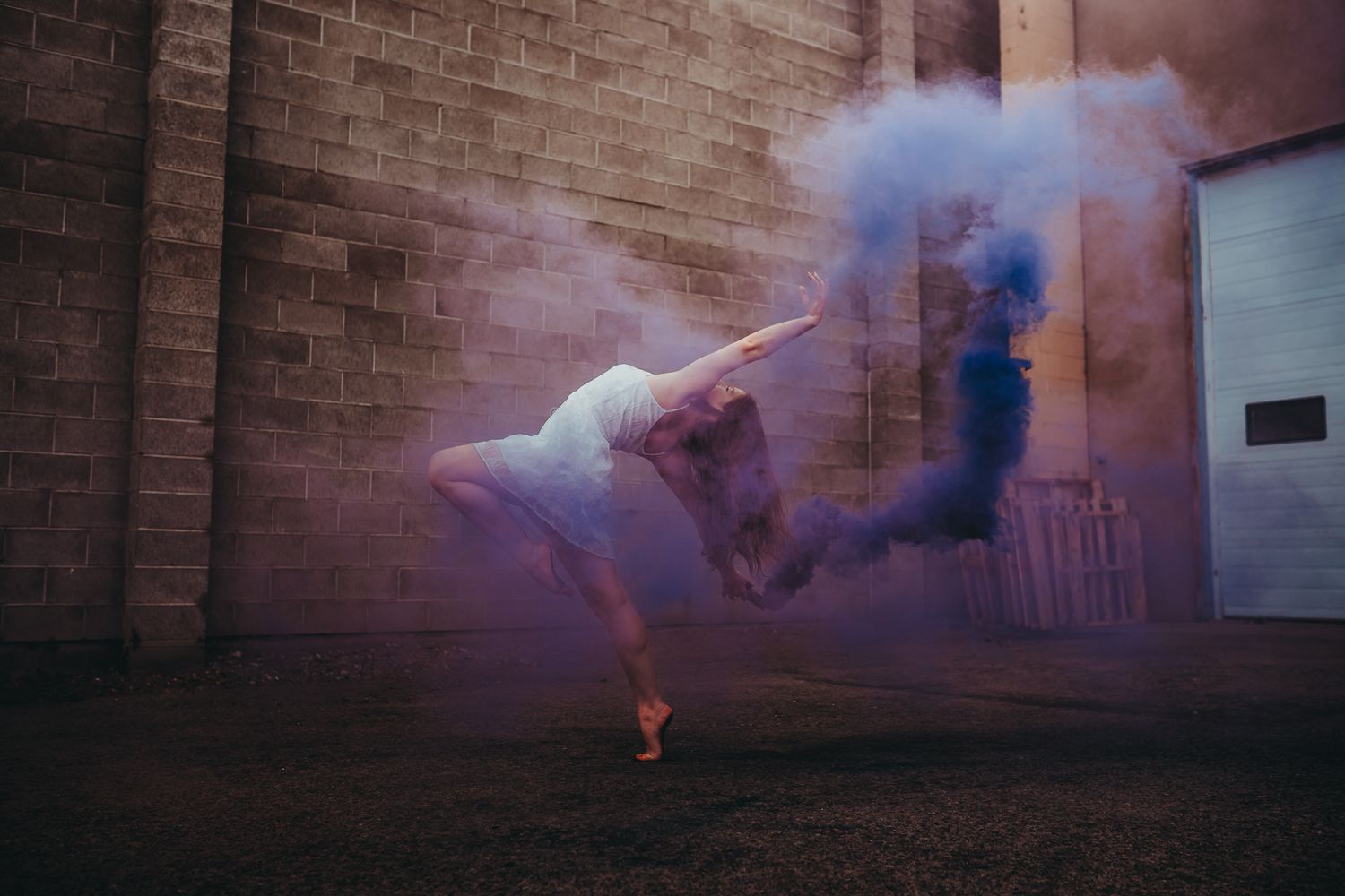 A dancer leaps gracefully through vibrant purple and blue smoke against a brick wall backdrop.