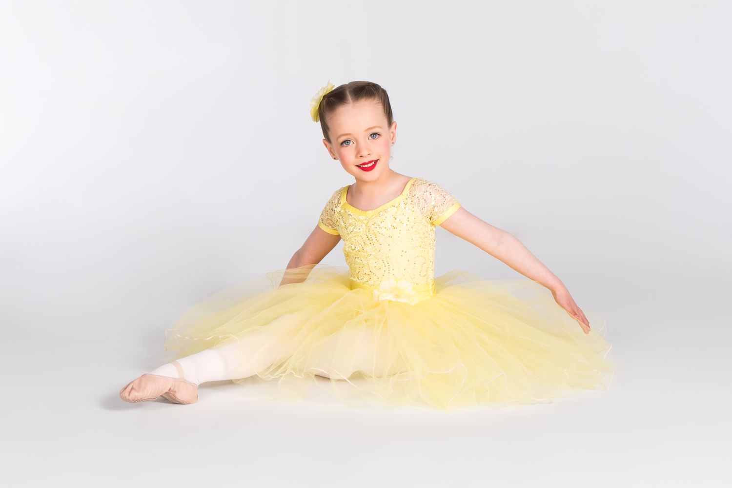 Dancer in yellow tulle dress poses gracefully in ballet position on white backdrop.