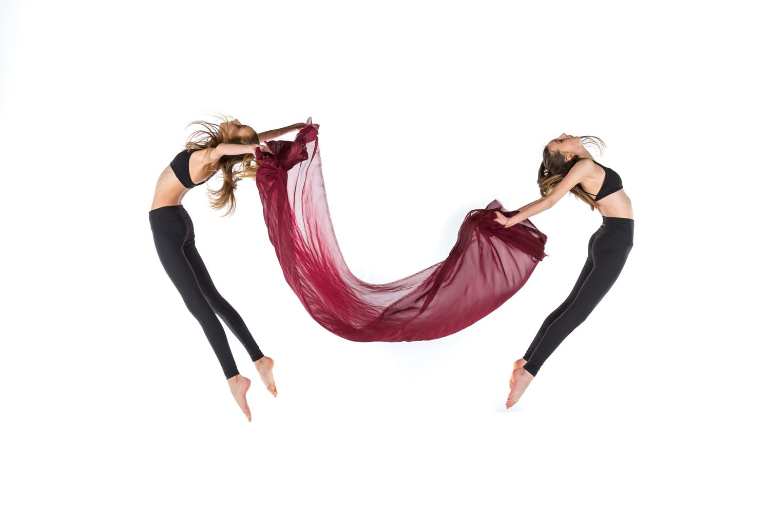 Two dancers in black leggings perform a graceful aerial pose while holding a flowing burgundy silk fabric between them.