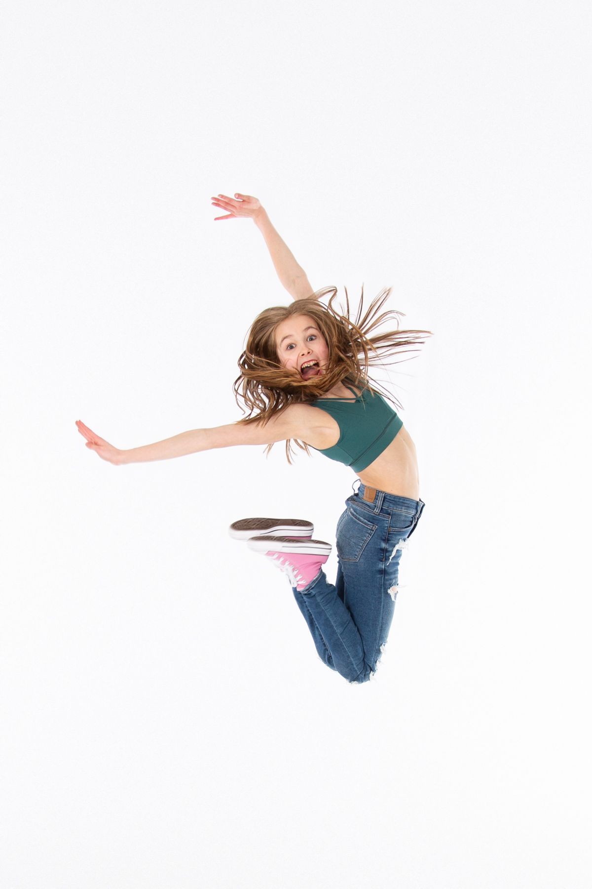 Young dancer in green crop top and jeans leaping joyfully with arms outstretched against white background.