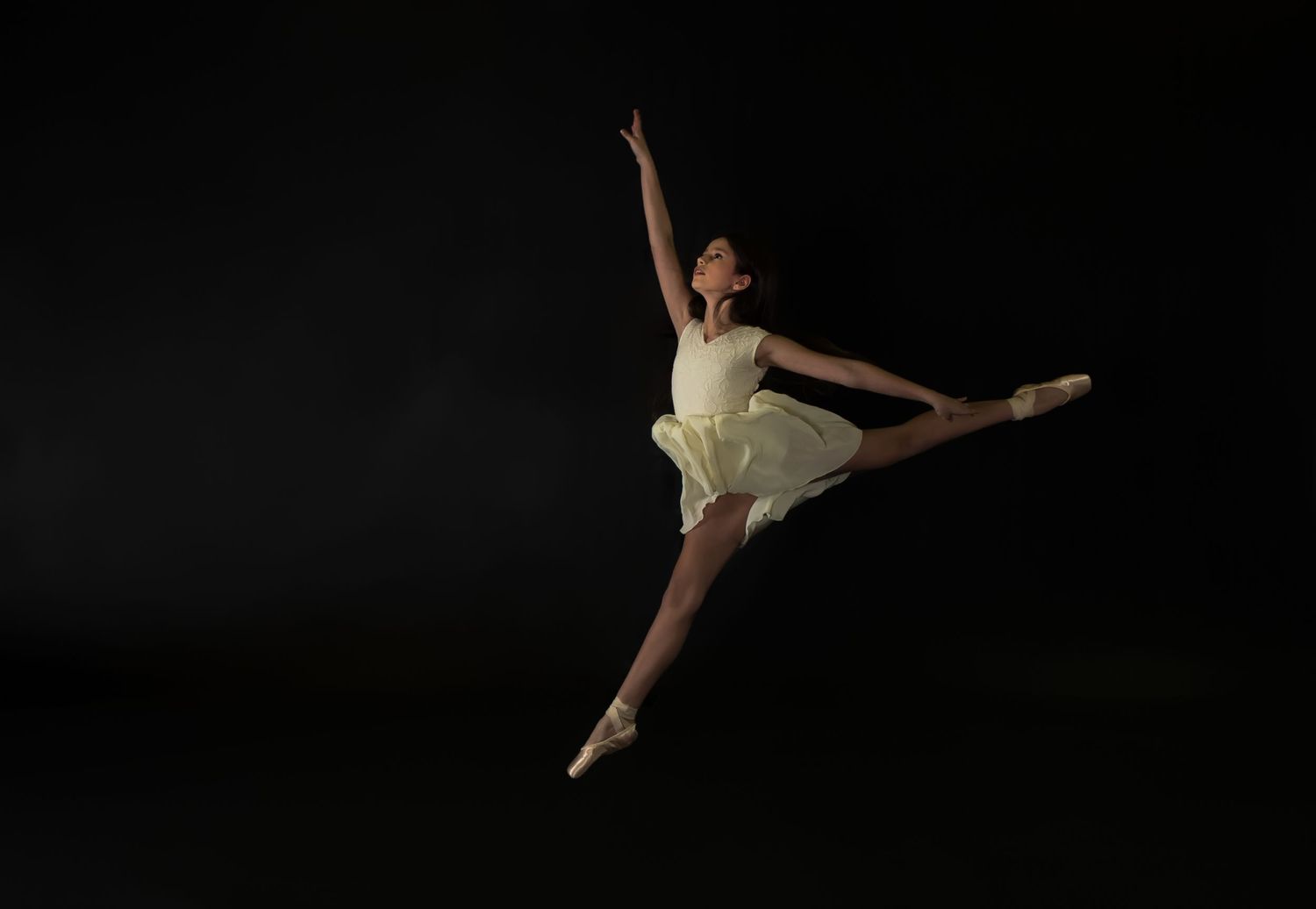 Ballet dancer in a light dress performs a graceful leap against a dark background.