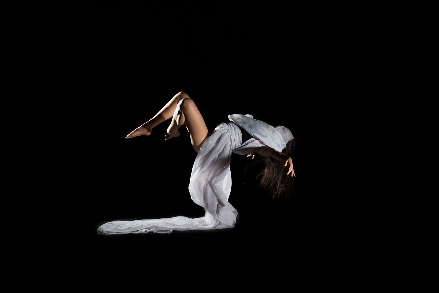 A dancer in white performs a graceful backbend pose against a black background during a contemporary dance performance.