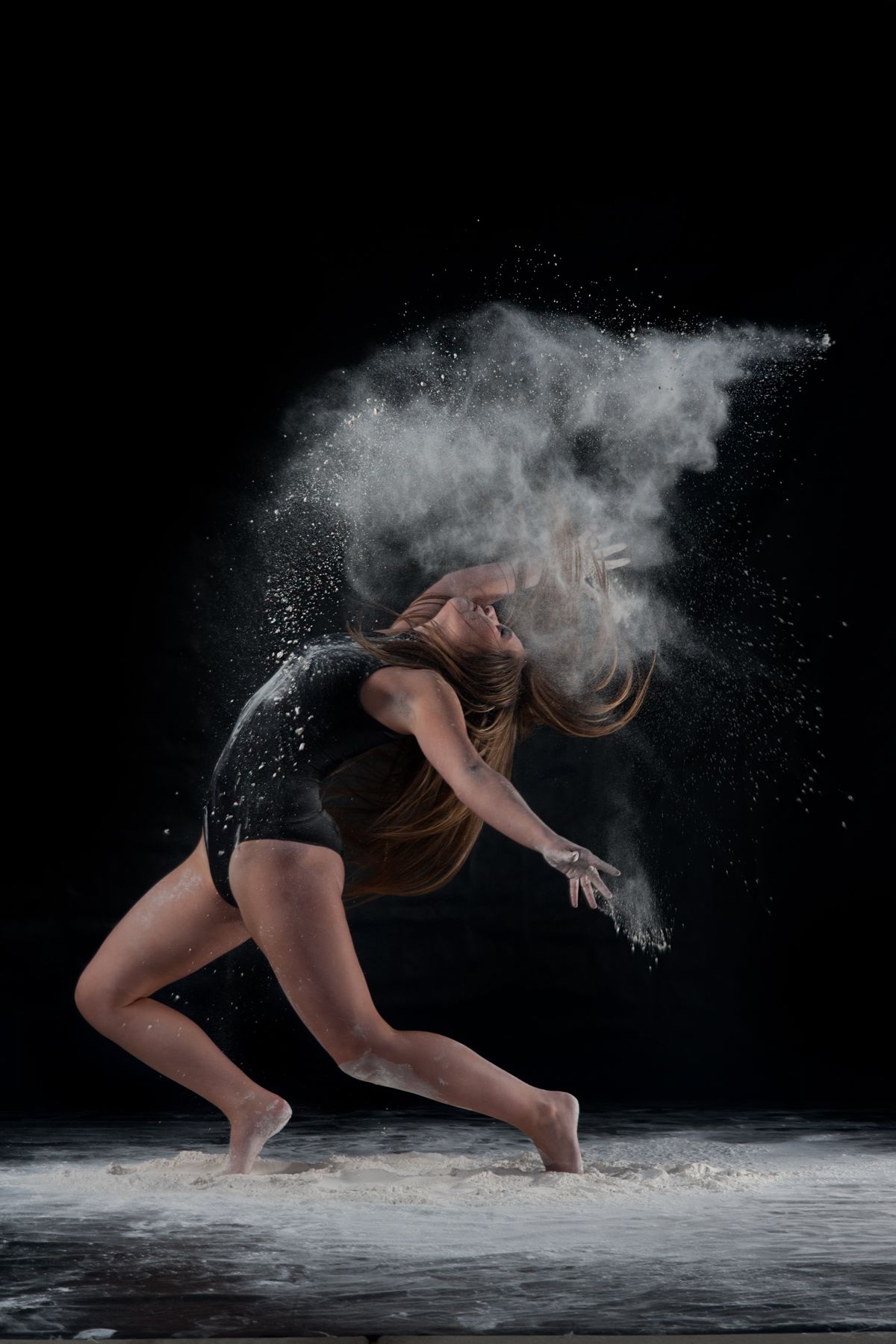 Dancer in black leotard performs dynamic movement with white powder creating ethereal effect against dark background.