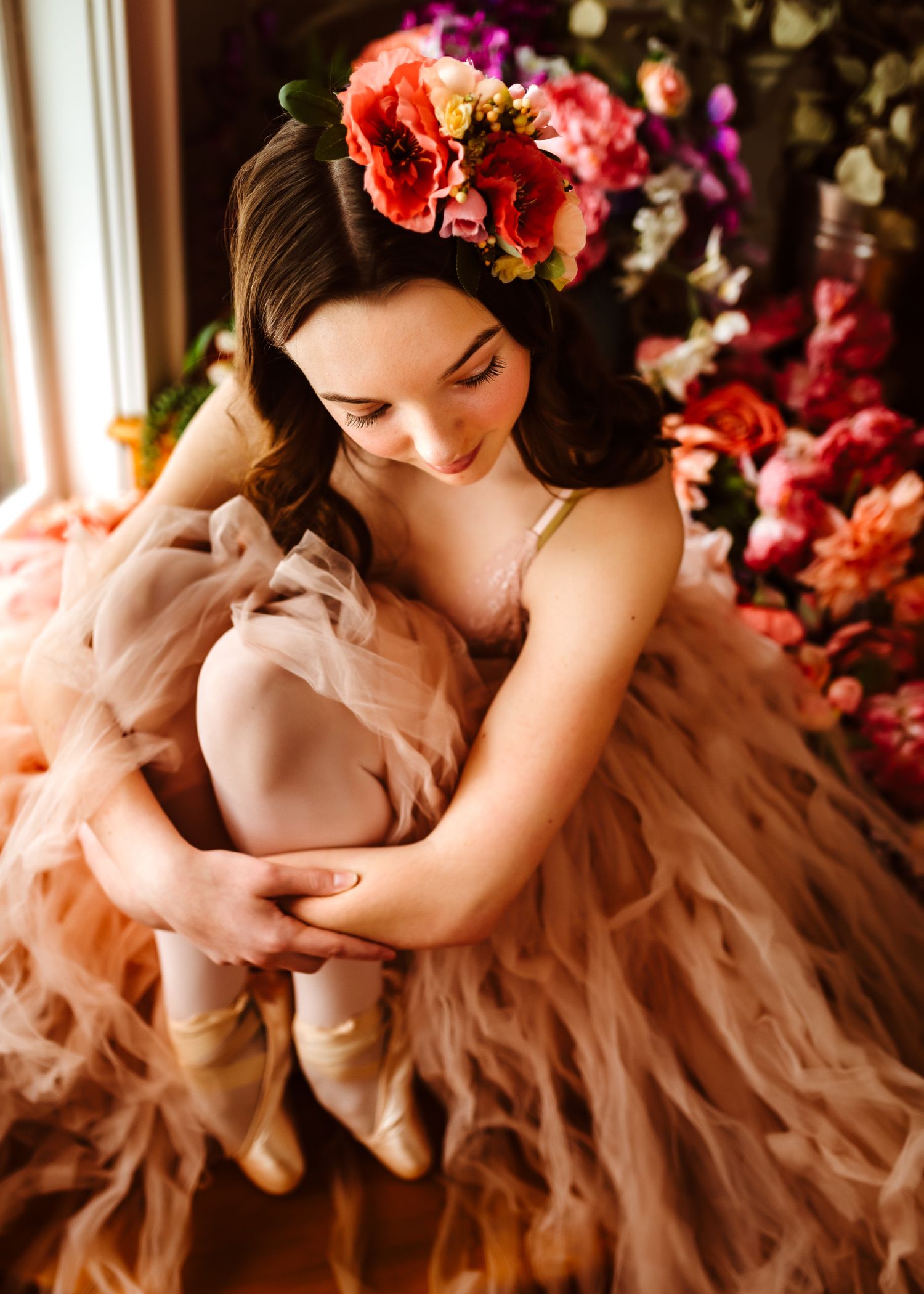 A person in a peach-colored gown wears a floral crown while sitting by a window surrounded by vibrant coral flowers.