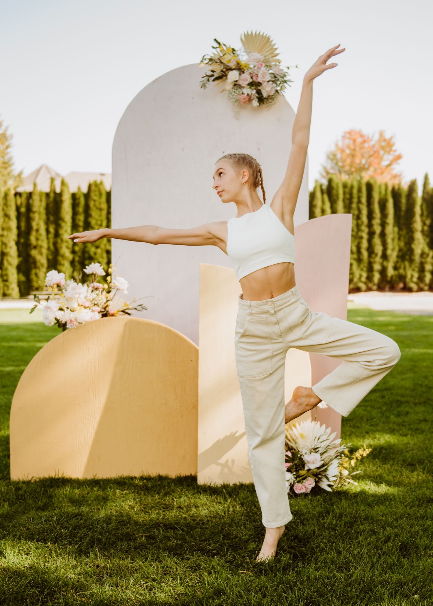 Modern dancer in white outfit performs elegant pose against geometric backdrop on sunny lawn.