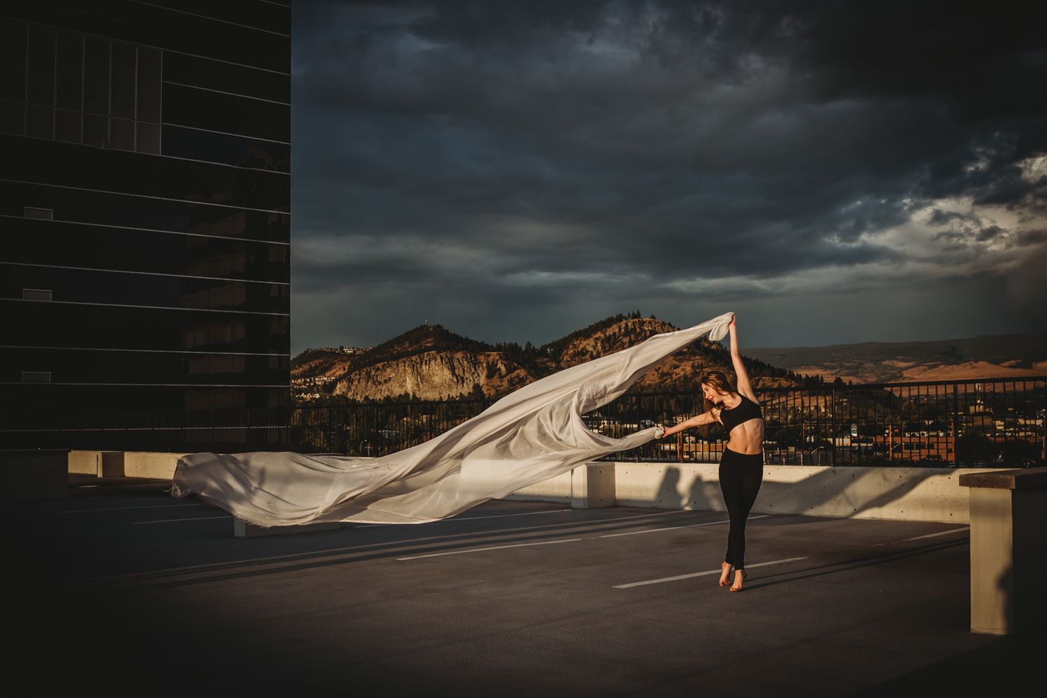 Dancer with flowing white fabric performs on rooftop against dramatic stormy mountain landscape.