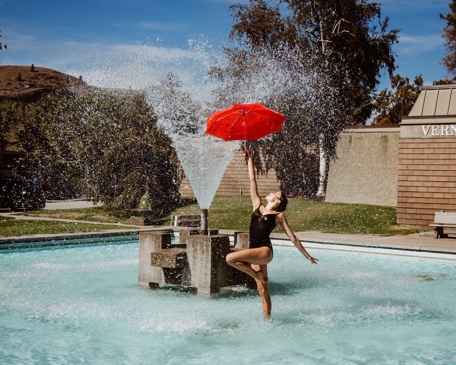 Someone with a red umbrella splashes in a fountain on a sunny day while water sprays up around them.