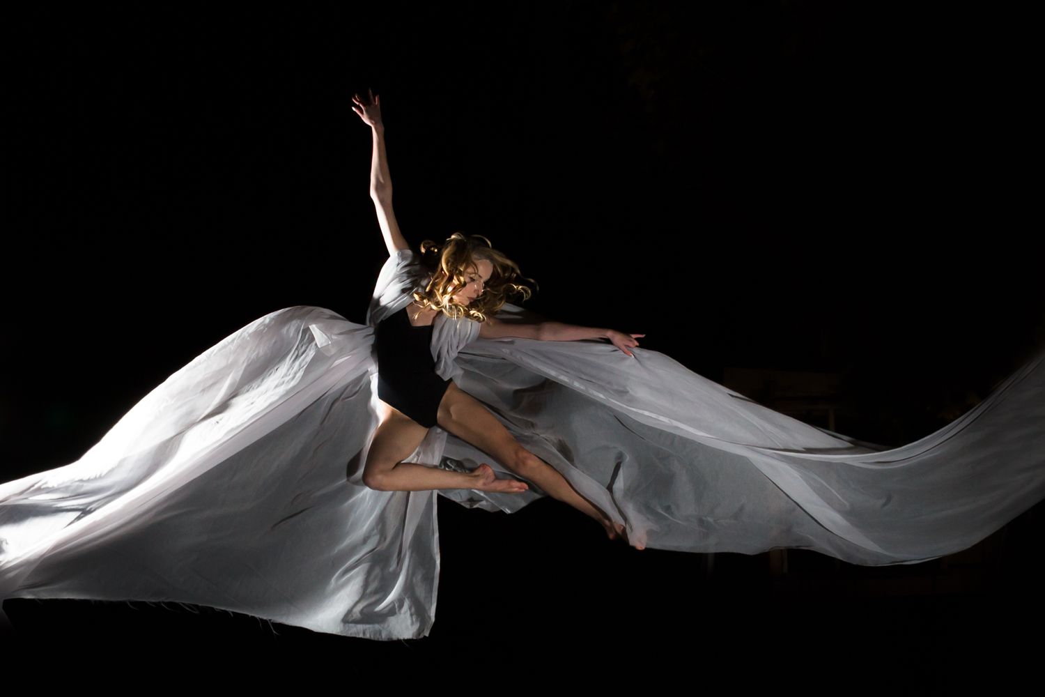A dancer leaps gracefully in flowing white fabric against a black background, creating an ethereal movement effect.