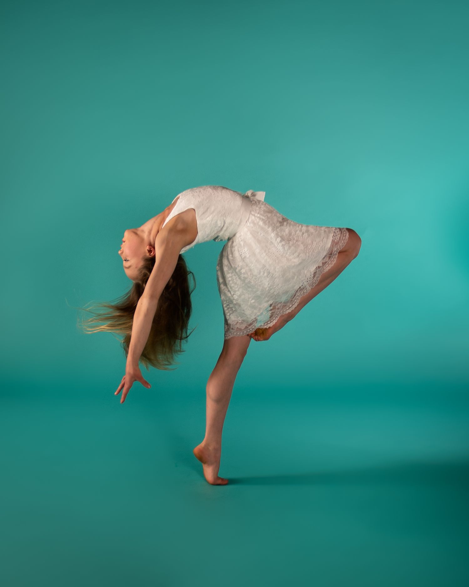 A graceful dancer in a white dress performs an elegant backbend against a teal background.