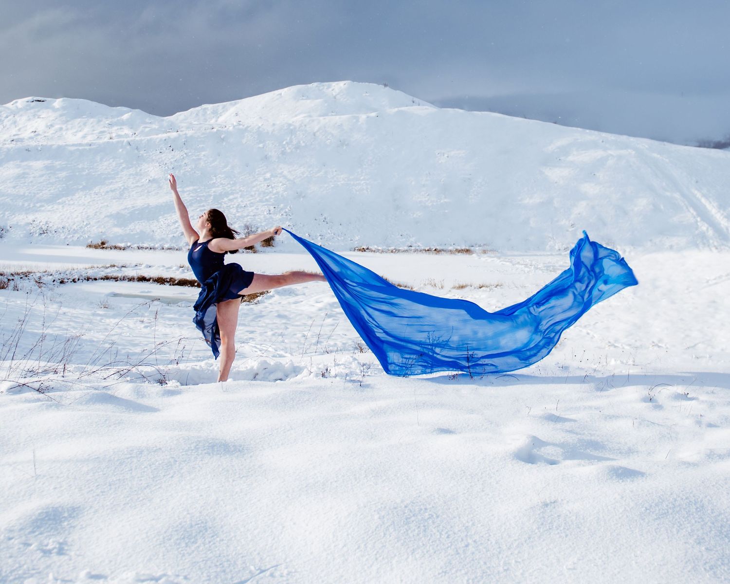 Dancer in black performs graceful pose on snow-covered landscape with flowing blue fabric against snowy mountains.