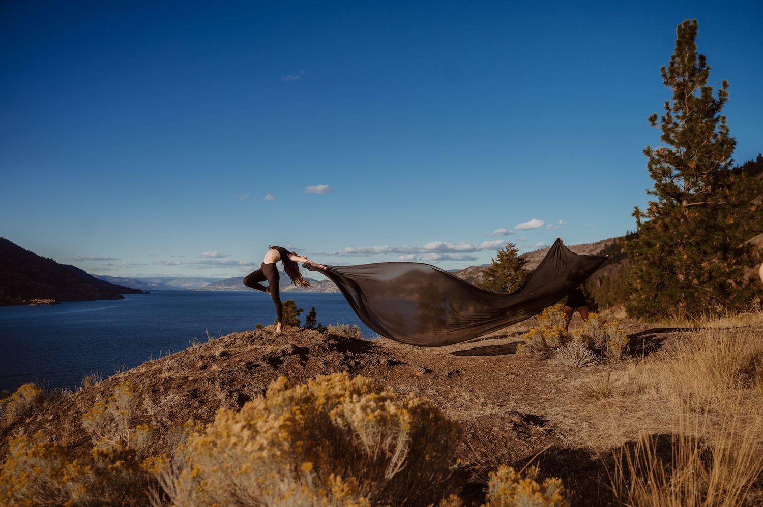 Silhouetted figure with flowing fabric stands on rocky cliff overlooking serene lake landscape at sunset.