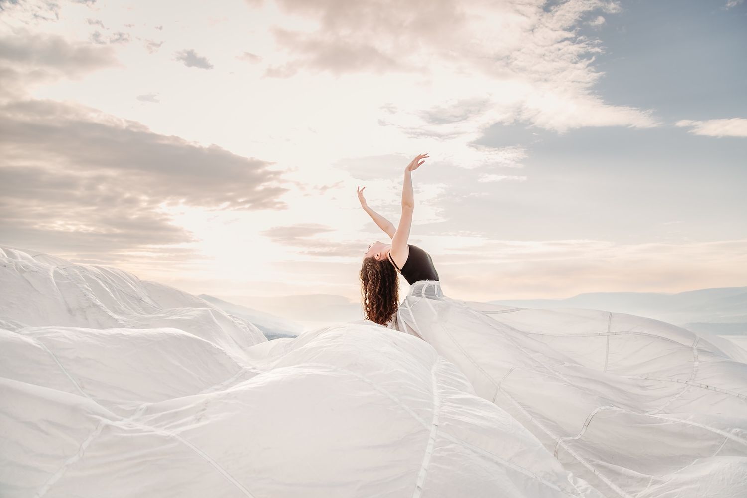 Silhouetted figure emerges from billowing white fabric against a soft sunset sky with mountains in the background.