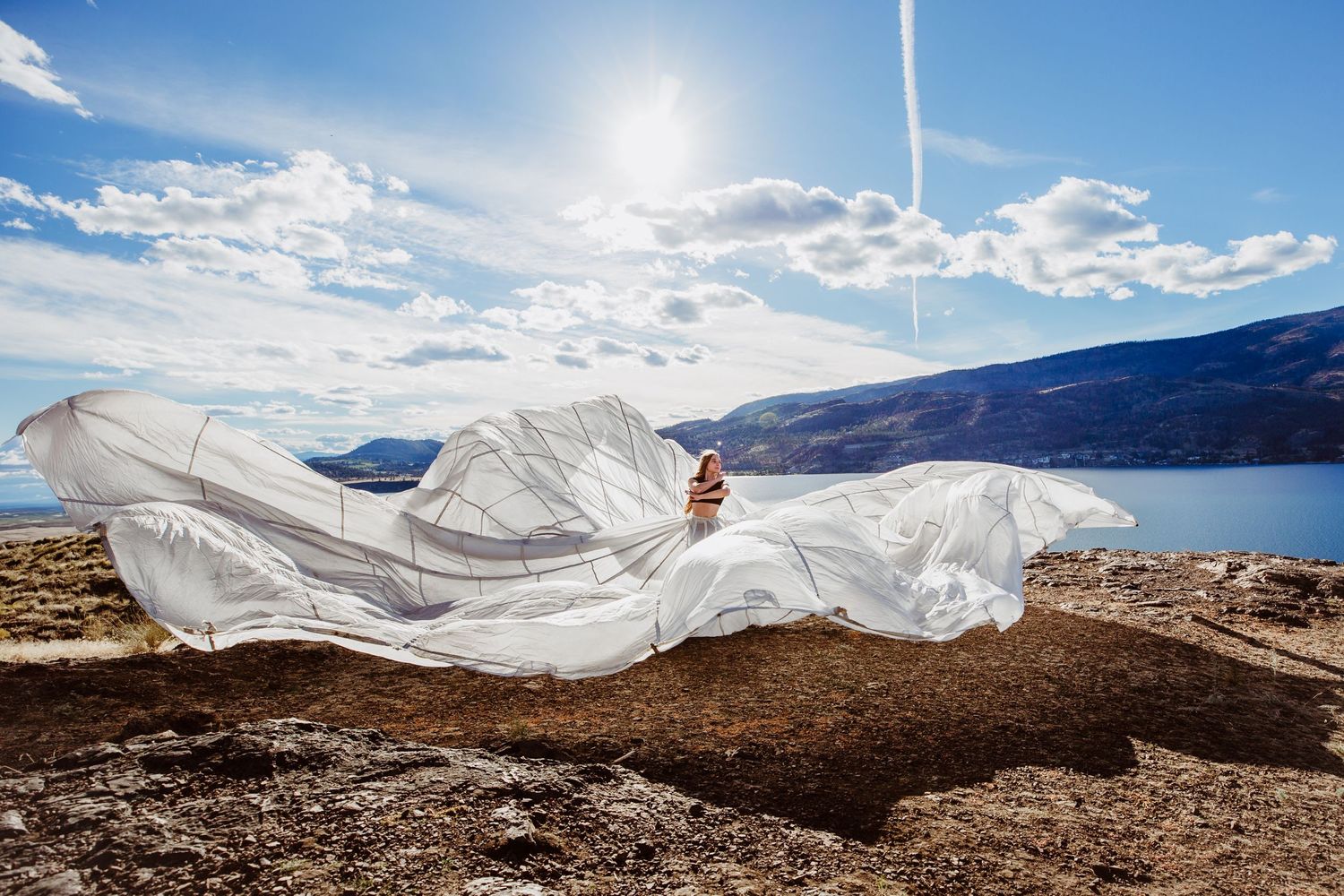 A flowing white wedding dress billows dramatically against a mountainous lake landscape on a sunny day.