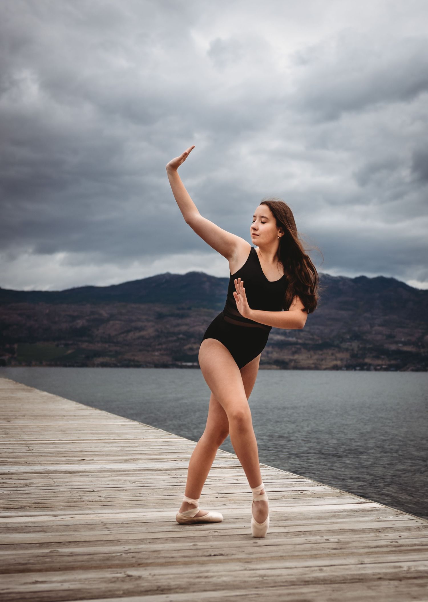 Graceful ballet dancer in black leotard performs en pointe on wooden dock overlooking misty mountain lake.