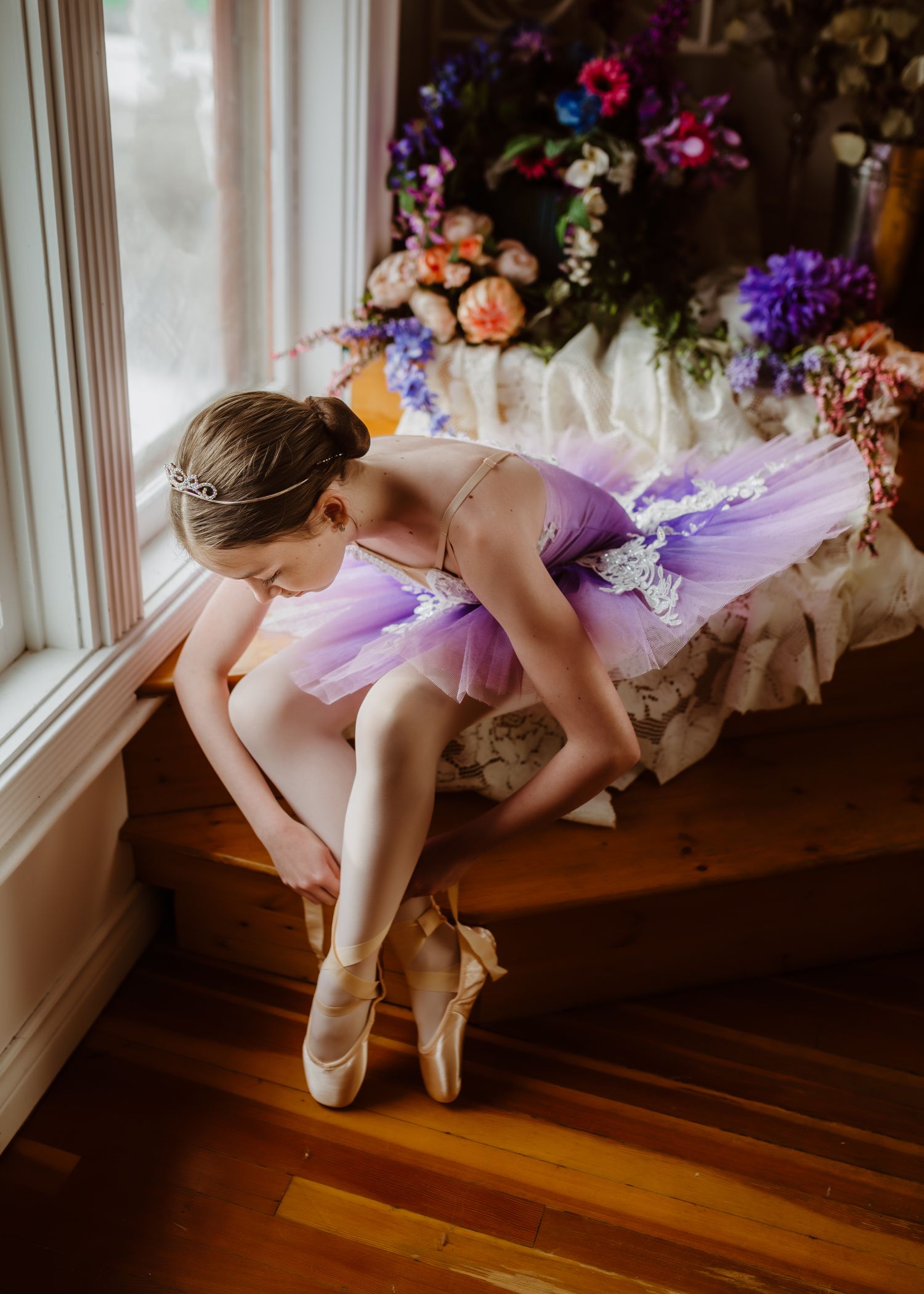 Young ballet dancer in purple tutu and pointe shoes leans gracefully by window with floral decorations.