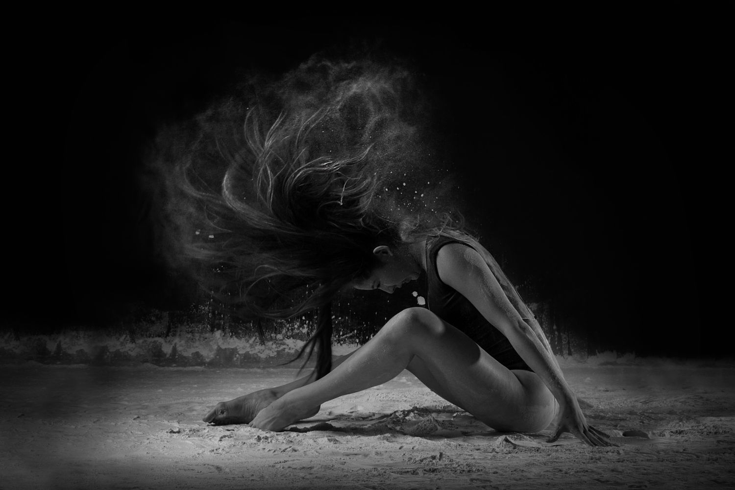 Artistic black and white photo of a dancer creating dramatic powder dust effect while performing on dark background.