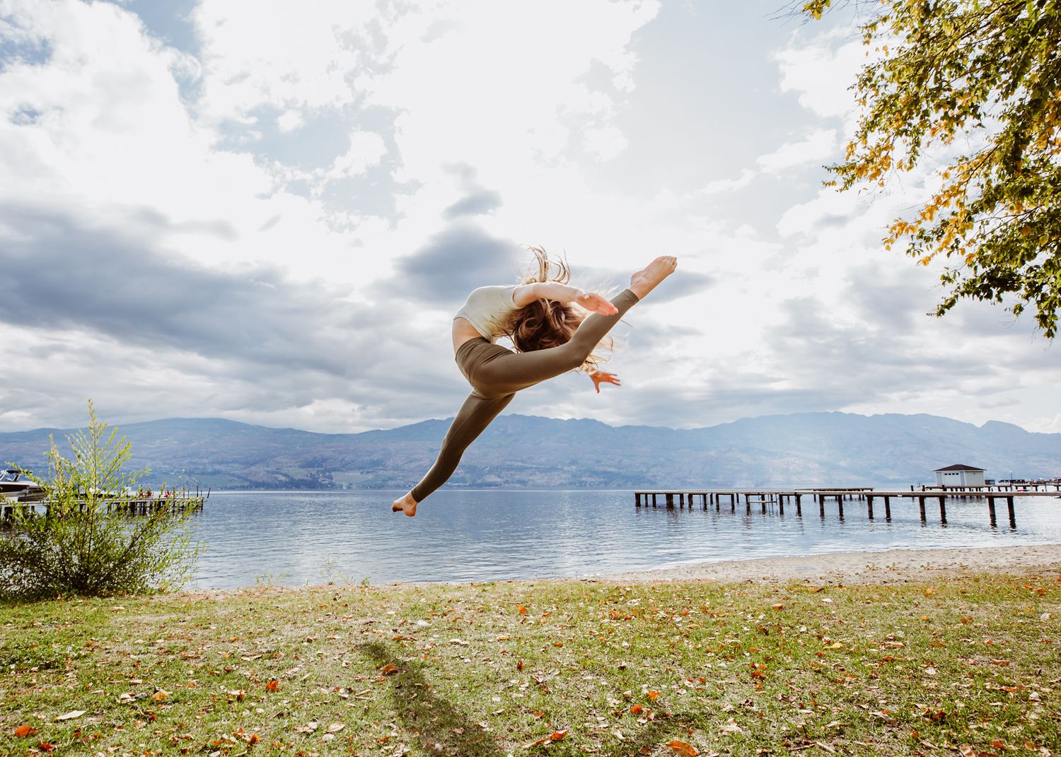 A dancer leaps gracefully against a scenic mountain lake backdrop with a wooden pier in the distance.