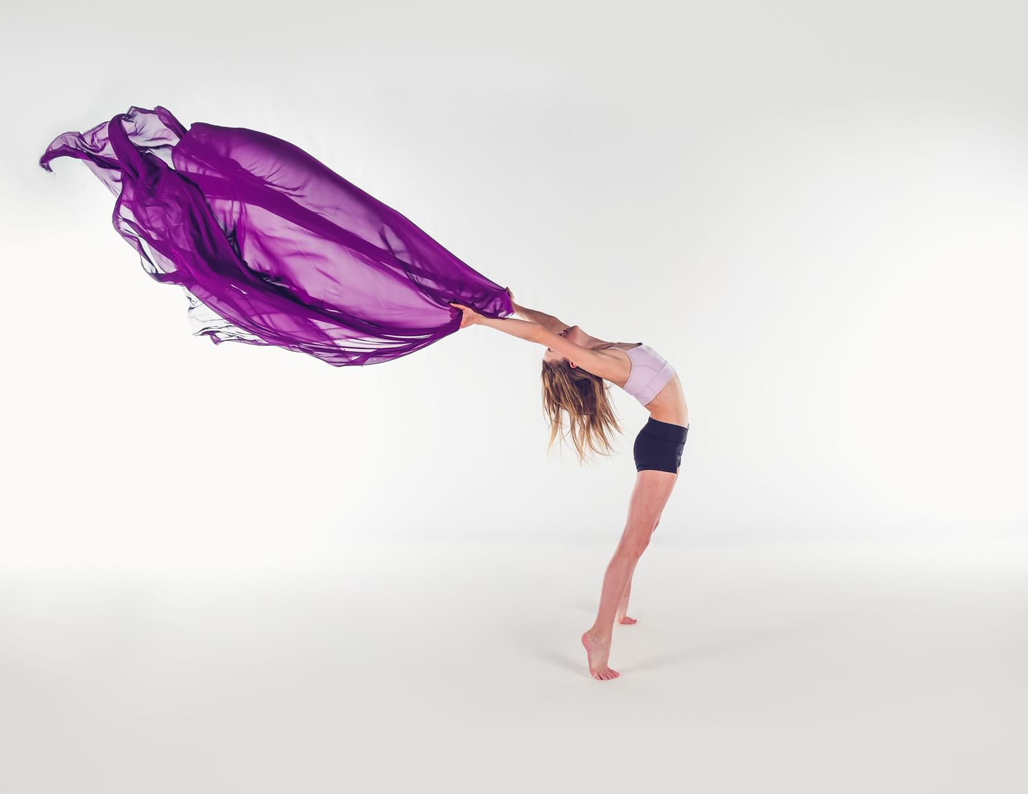 A graceful dancer bends backwards with a flowing purple silk fabric billowing dramatically against a white background.