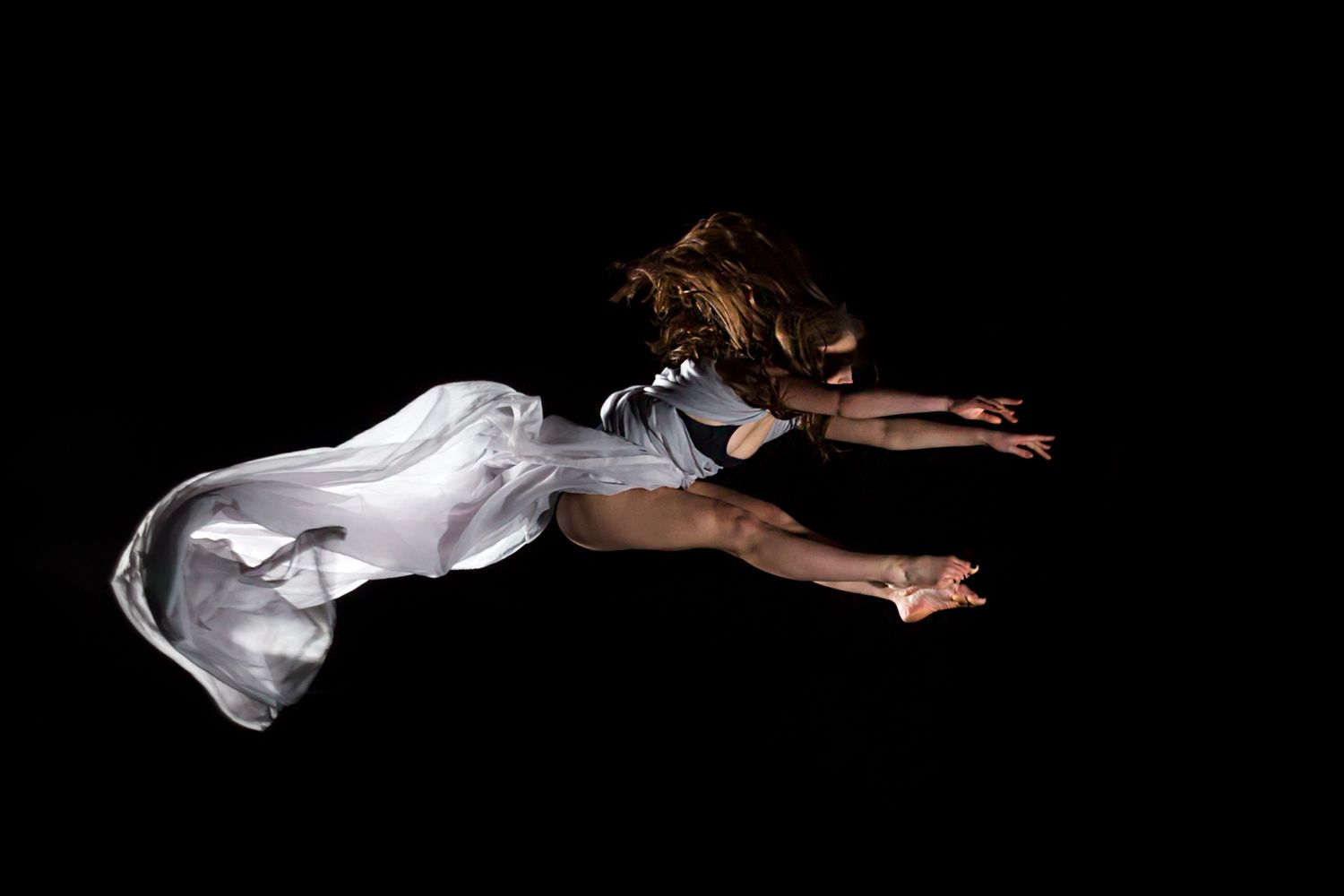 Graceful dancer soars horizontally through air with billowing white fabric in dramatic lighting.