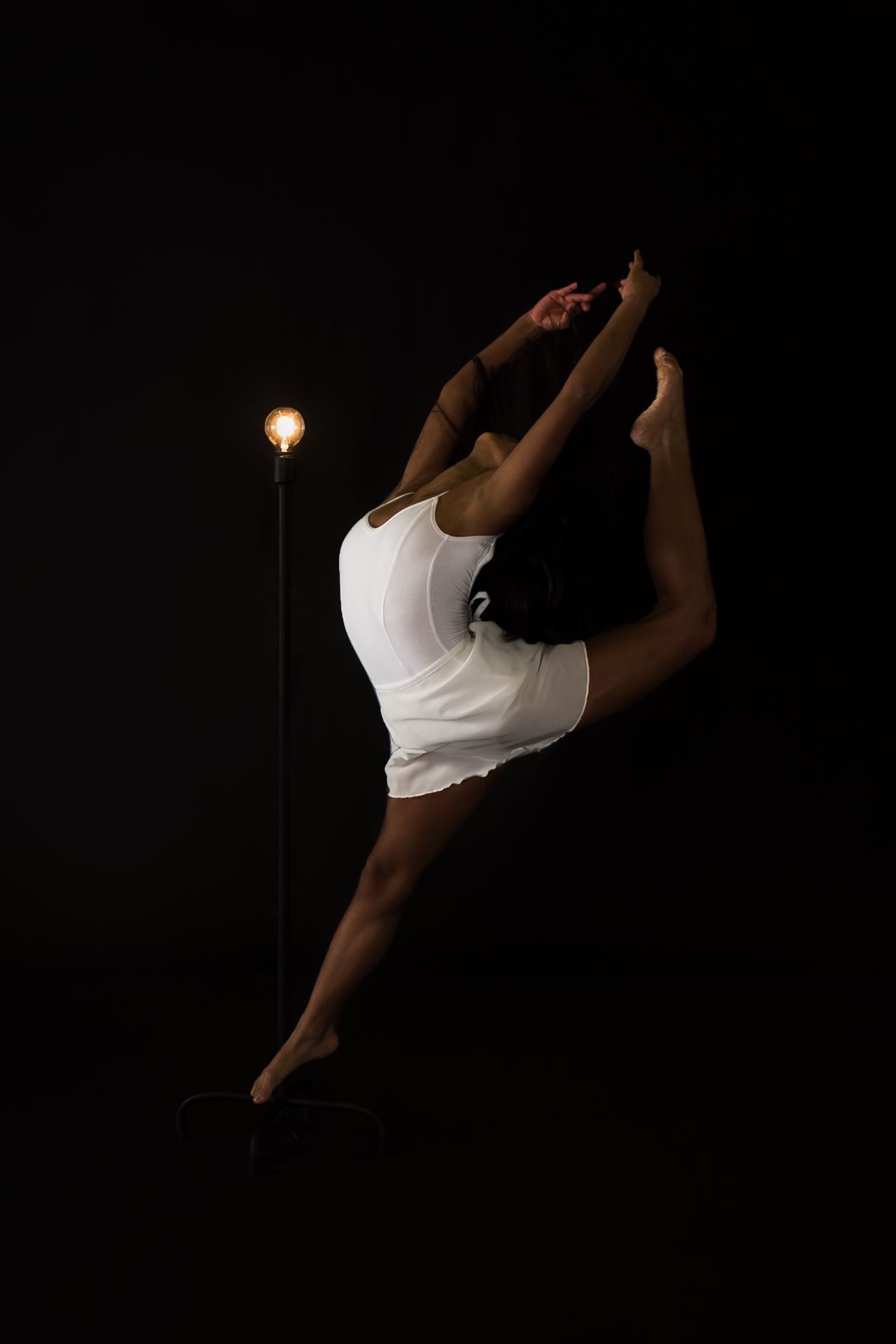 A dancer in white performs a graceful leap against a dark backdrop with a single glowing light in the background.