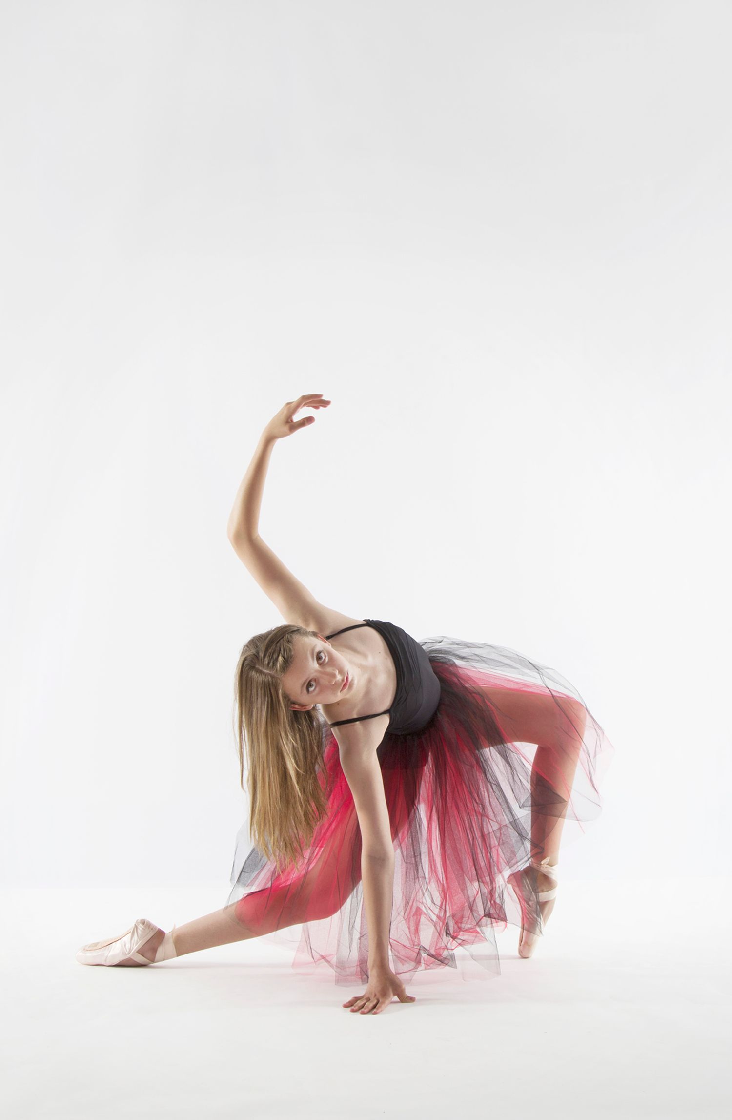 A ballet dancer in a pink tulle skirt performs an elegant bending pose on a white backdrop.