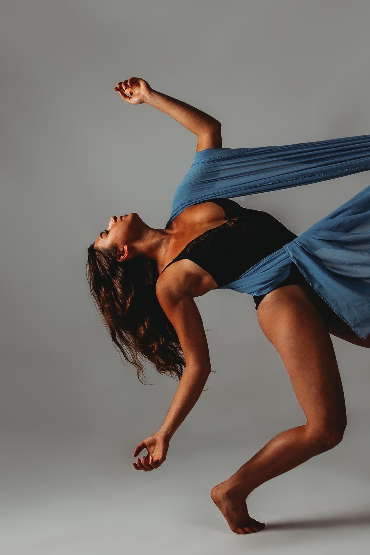 Contemporary dancer in blue dress performing emotive backbend against gray backdrop.