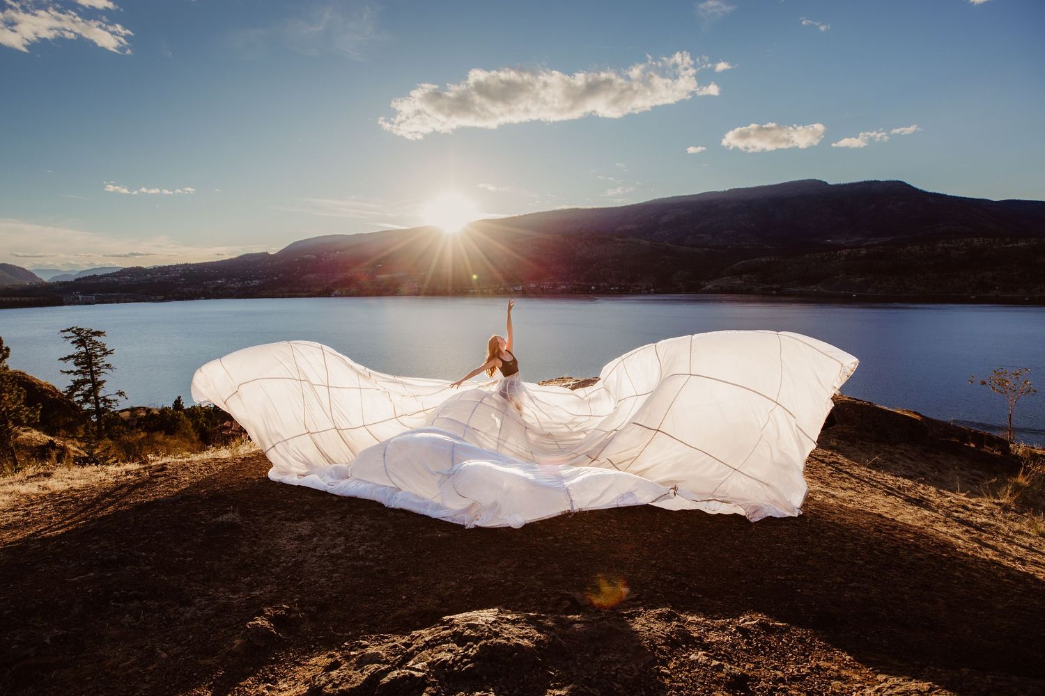 A flowing white dress billows in the wind against a backdrop of a sunset over a mountain lake.