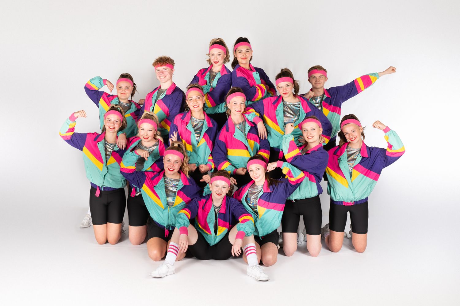 A dance group poses together in colorful geometric patterned uniforms with bright pink, teal and black elements.