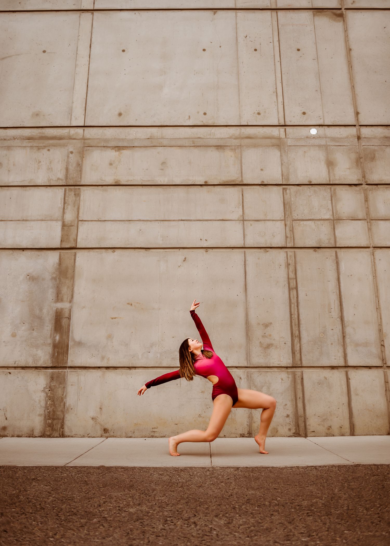 A dancer in a red leotard performs a dynamic leap against a concrete wall background.