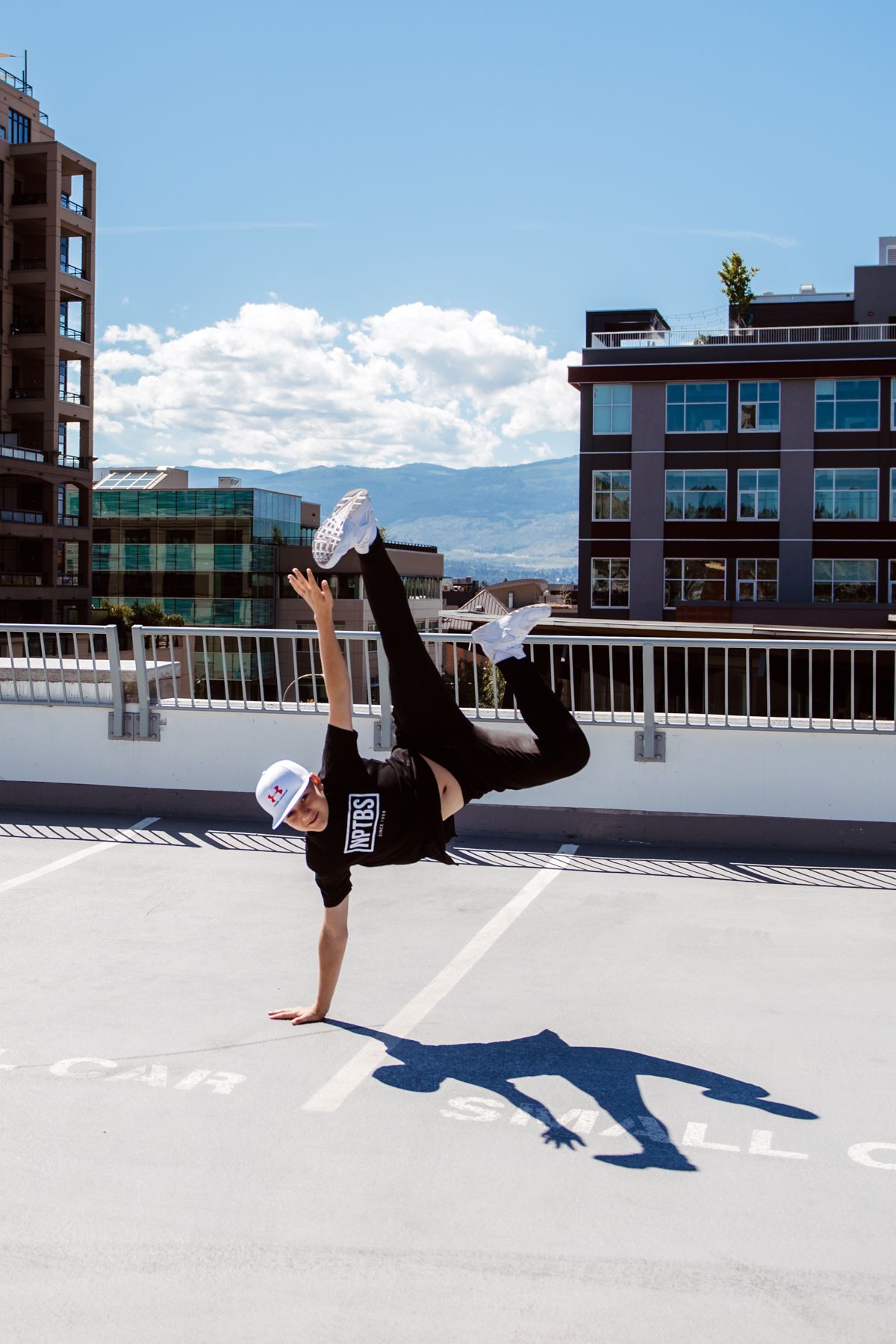A dancer performs a one-handed handstand on a rooftop with city buildings and mountains in the background.
