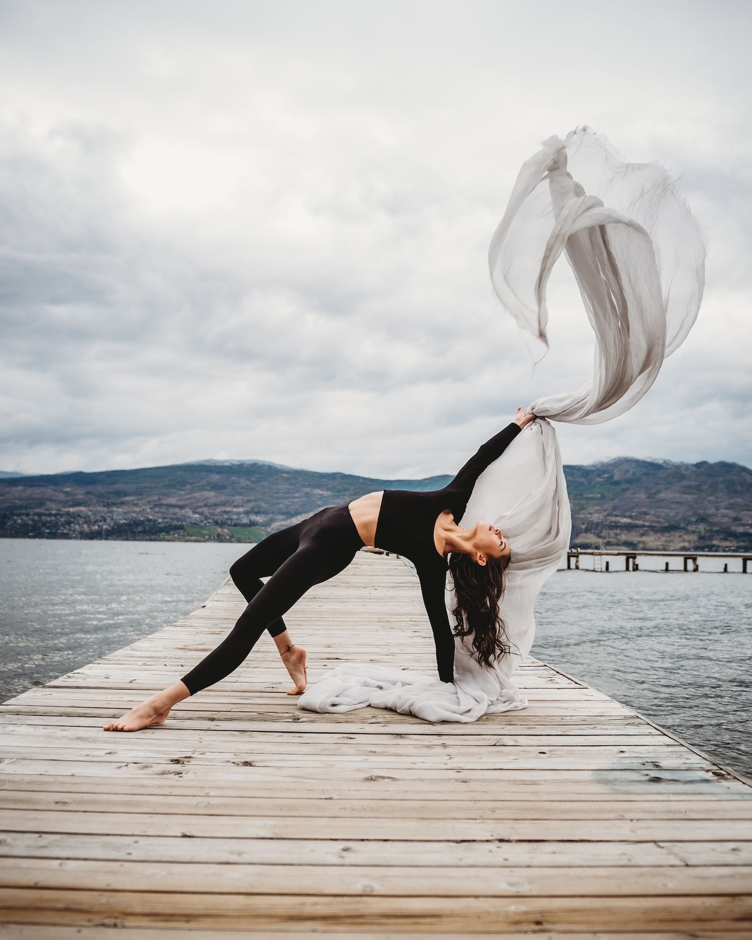 Dancer poses on wooden pier with flowing fabric against mountain lake backdrop.