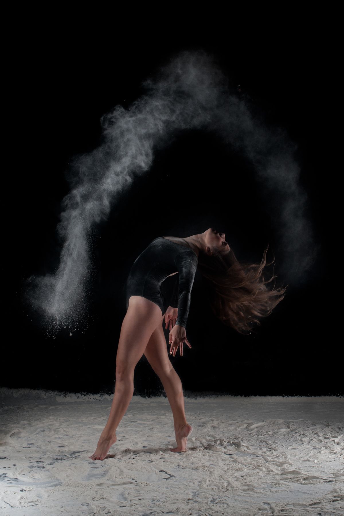Dynamic dancer in black leotard performs dramatic backbend as white powder creates arc of dust in dark studio.