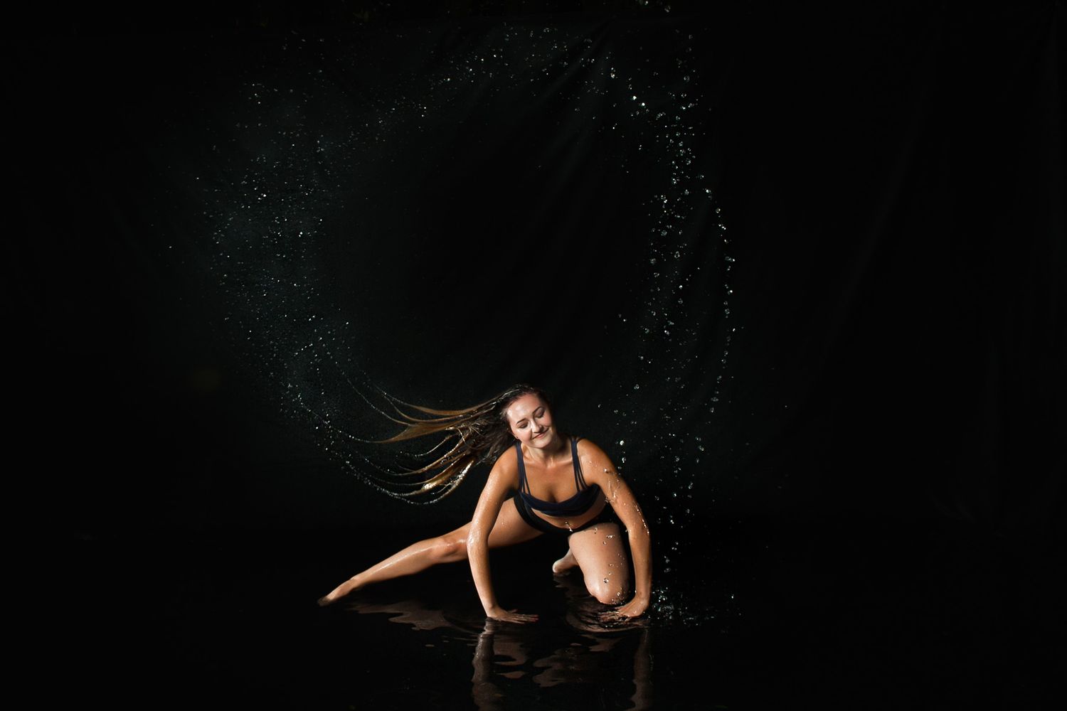 Athletic dancer in black creates dramatic water splash effect during dynamic performance in dark studio setting.
