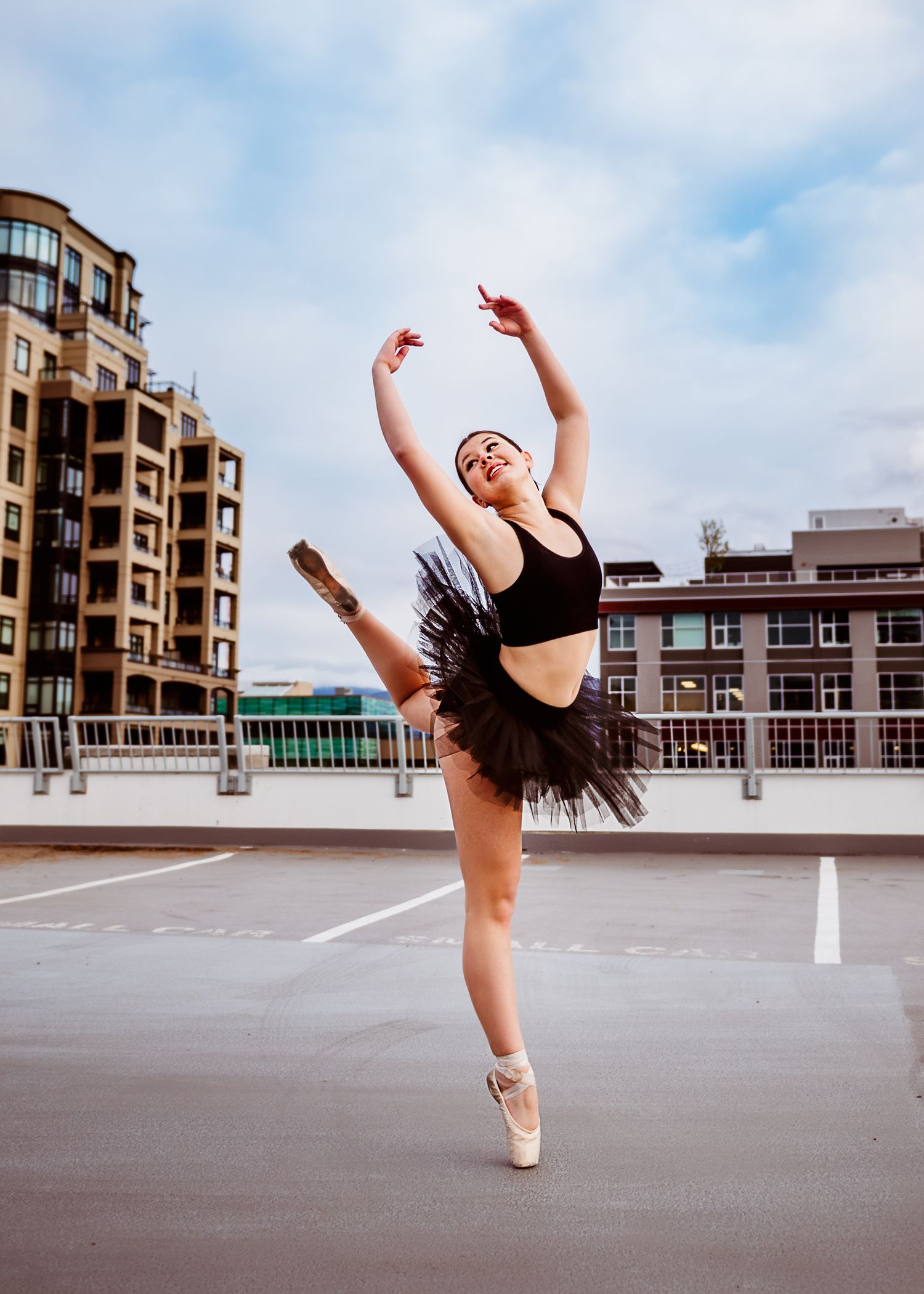 A ballet dancer in pointe shoes and black tutu performs an arabesque pose on a rooftop with city buildings in the background.