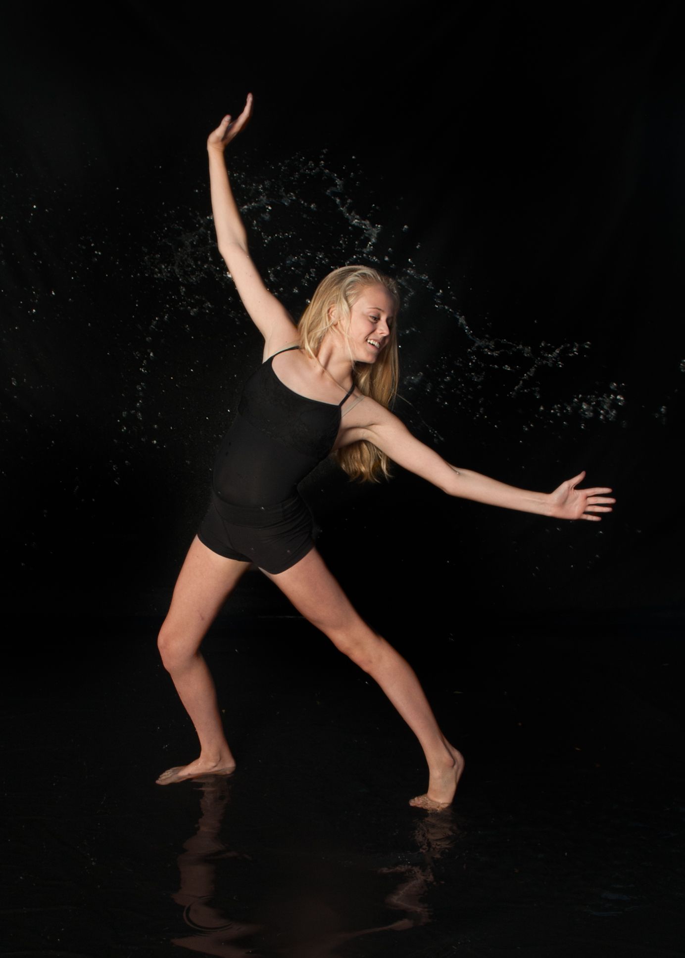 Contemporary dancer in black outfit performing expressive movement with water splashes on dark stage.