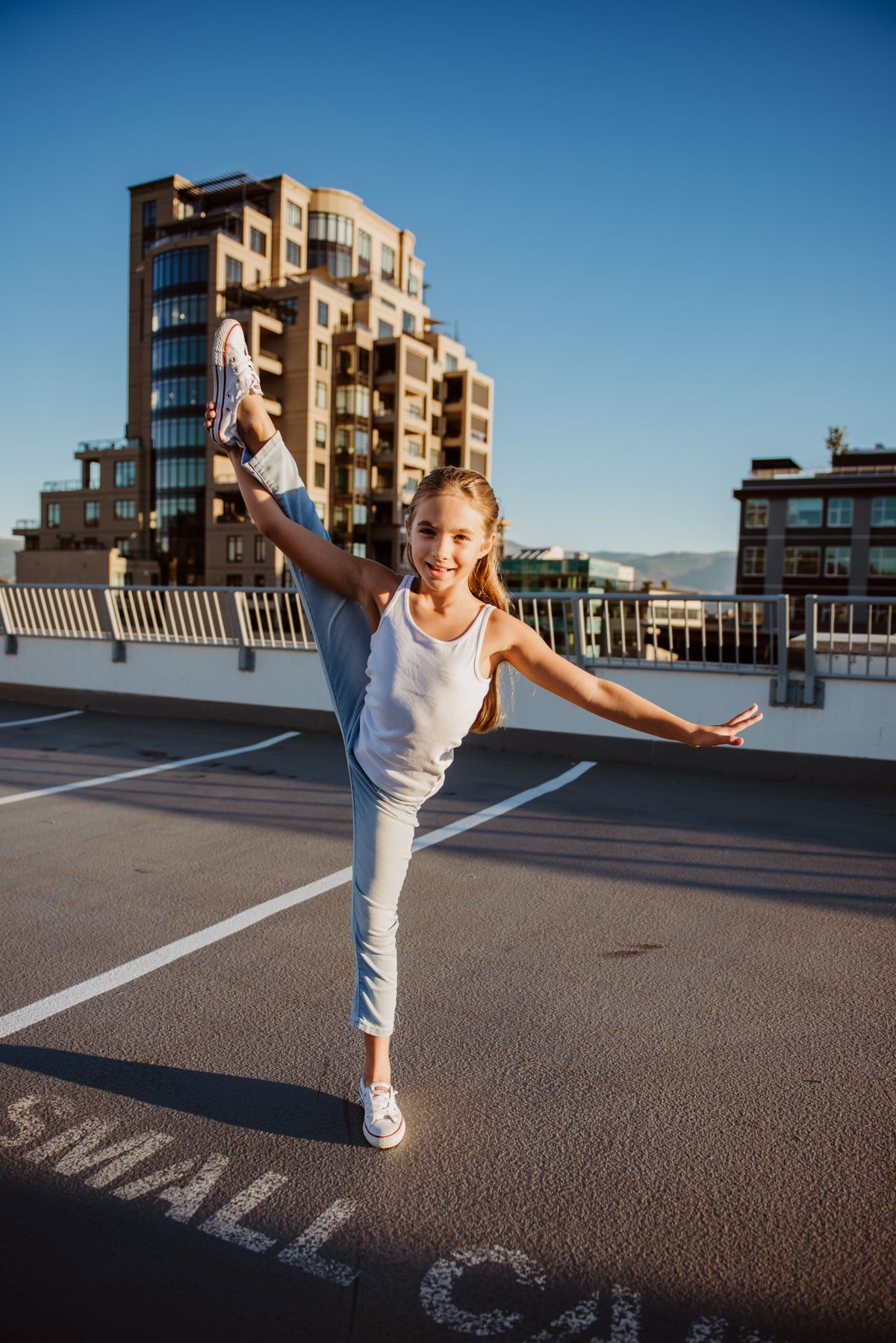 Young dancer strikes an athletic pose on a rooftop parking area with urban buildings in the background.