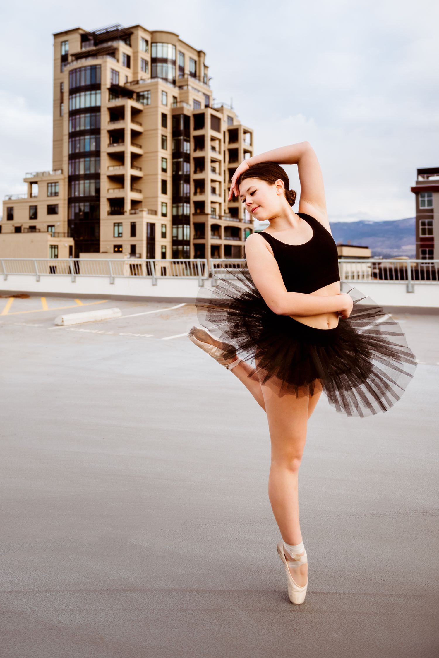 A ballet dancer in a black tutu performs an elegant pose on a rooftop with urban architecture in the background.