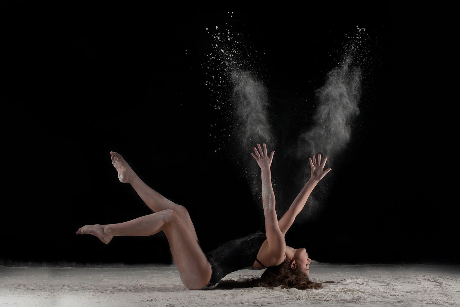 Dancer laying in white powder creates dramatic cloud effect during artistic performance on dark stage.
