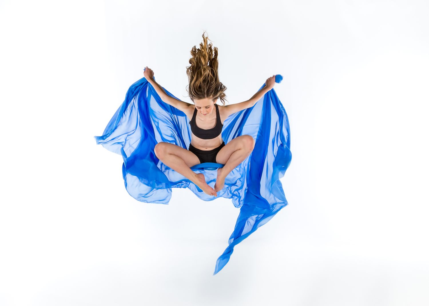 A dancer in black leotard leaps gracefully with flowing blue fabric against white background.