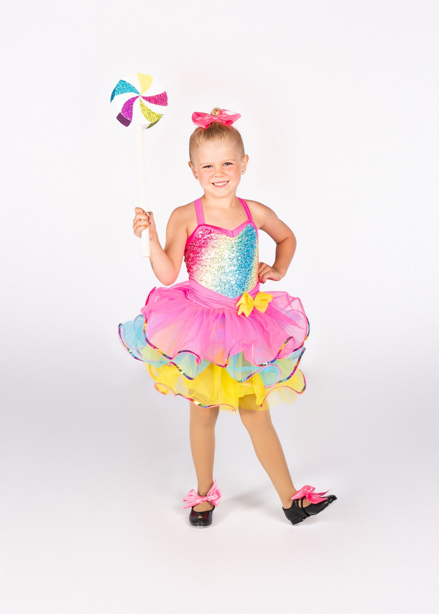 Dancer in sparkly rainbow costume with pink and yellow tutu performs with colorful ball against white backdrop.