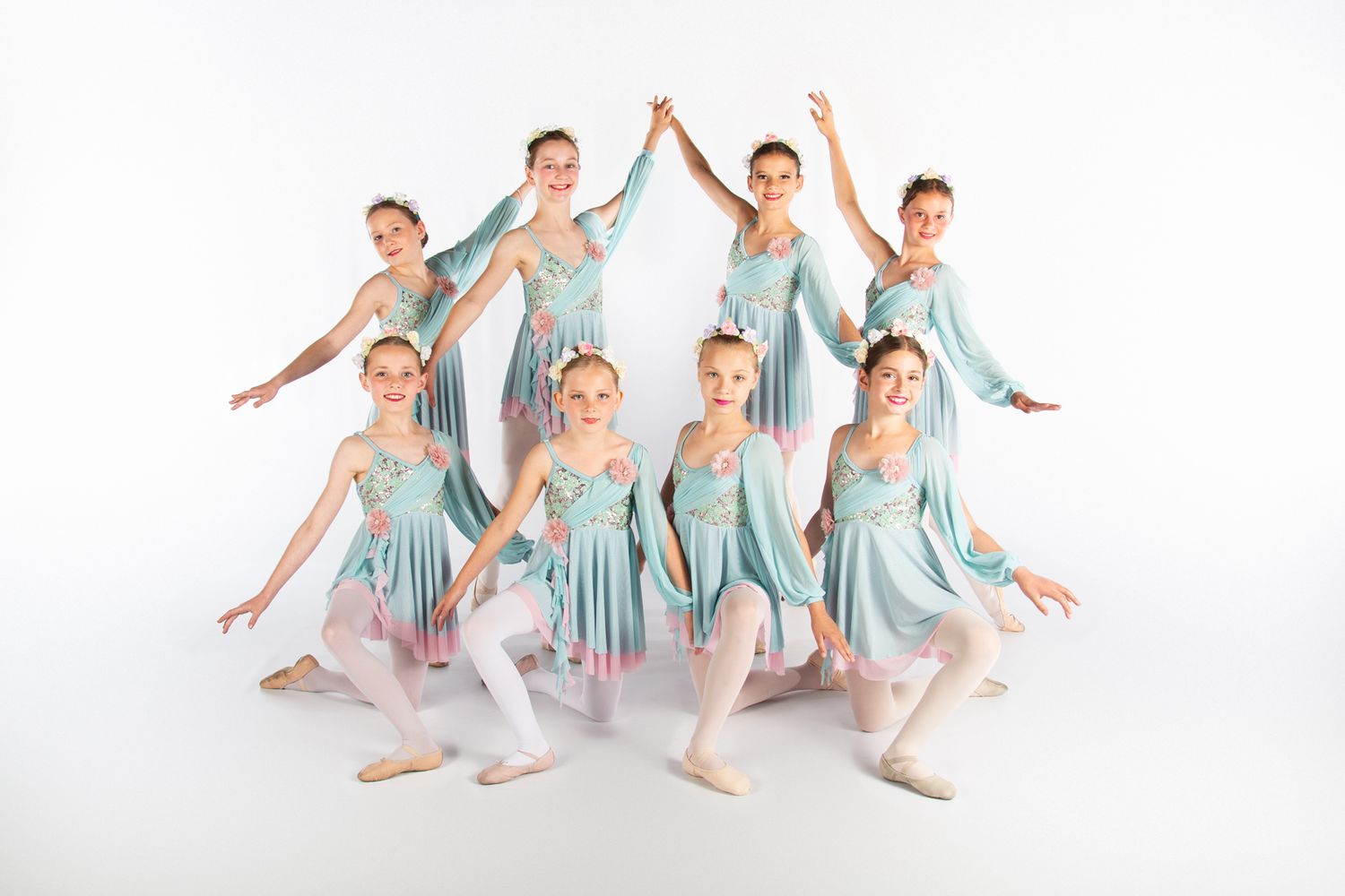 Dance students in light blue dresses perform graceful ballet poses together in formation against white background.