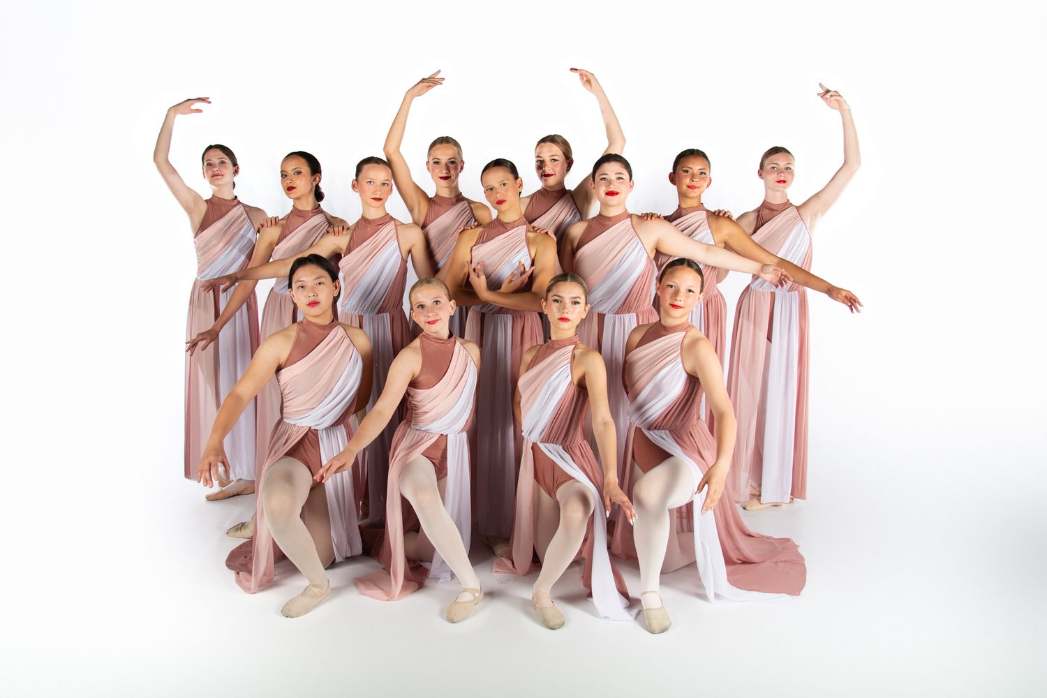 Ballet dancers in flowing beige costumes strike graceful poses together against a white backdrop during a group performance.