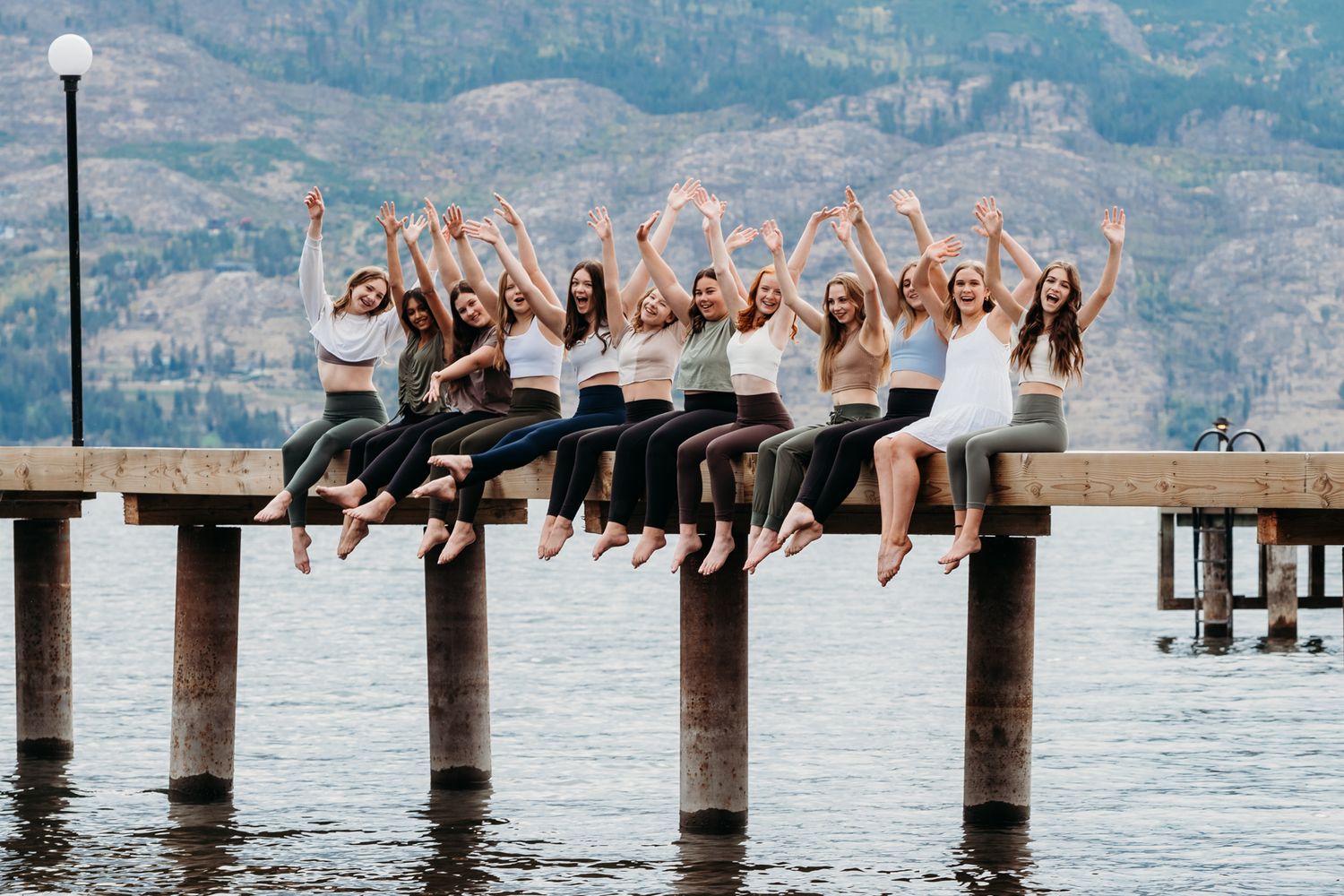 A group of friends in black dresses sitting together with raised arms on a wooden pier over a lake with mountains in background.