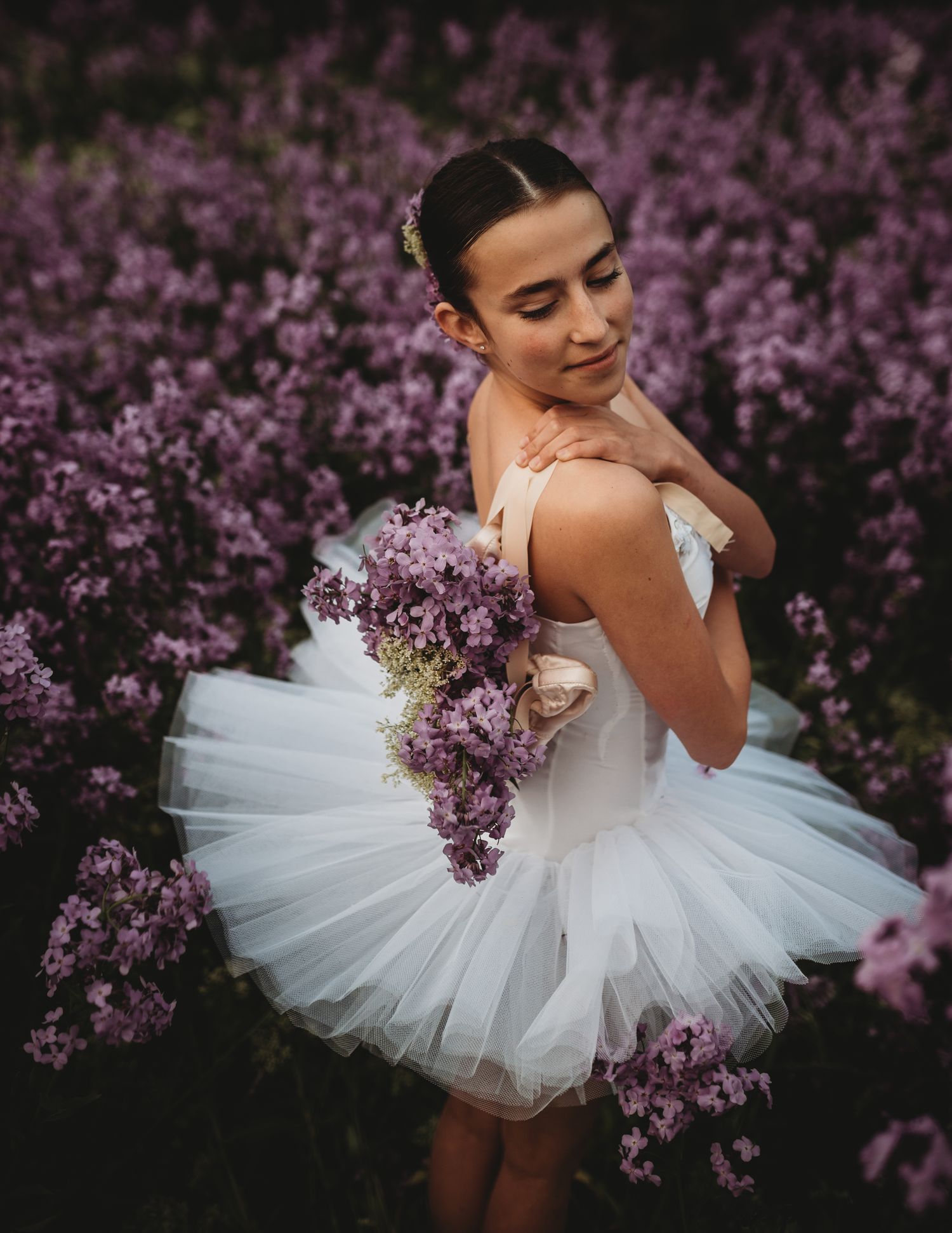 A ballerina in a white tutu poses gracefully among blooming purple lilac bushes while holding a floral bouquet.