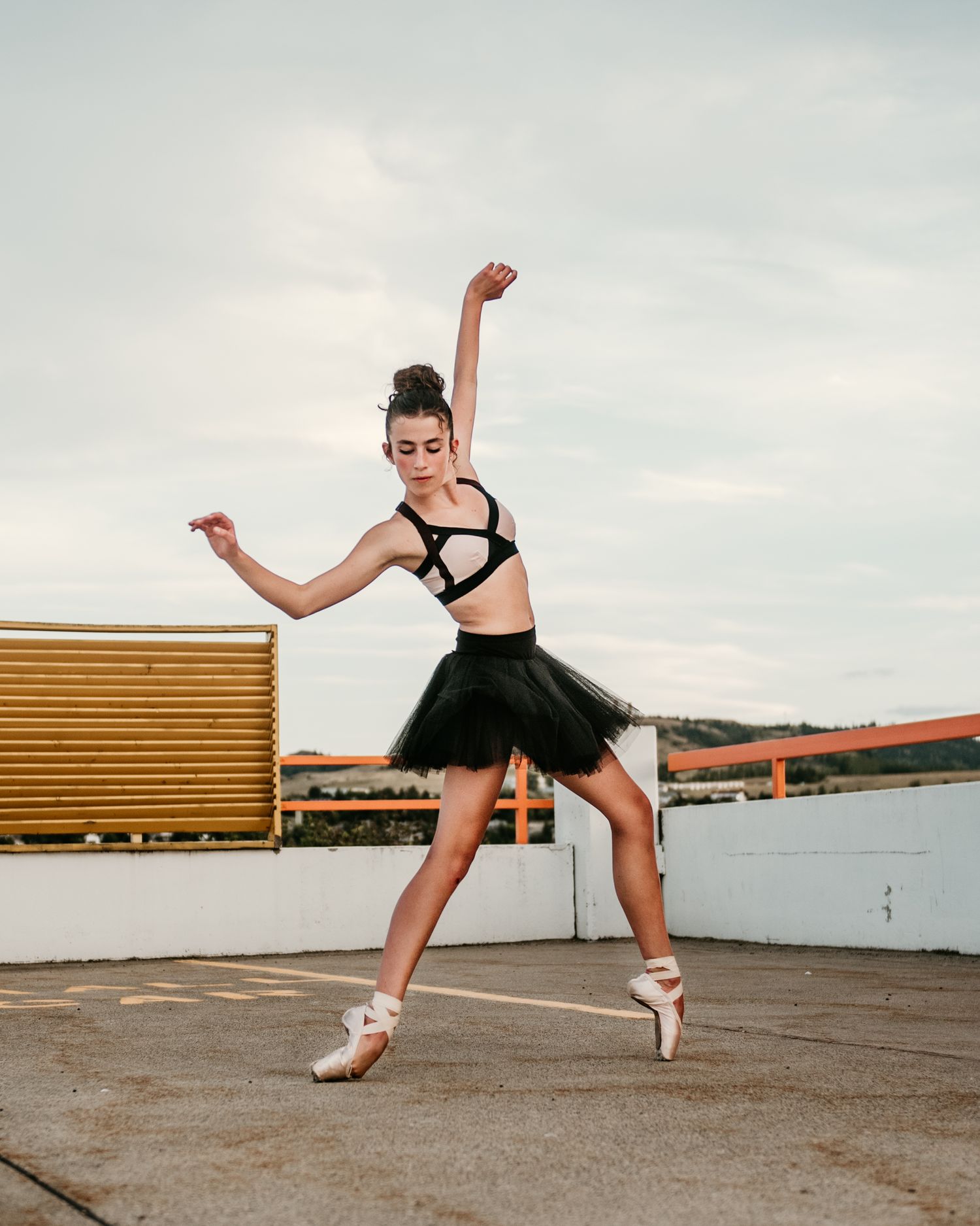 A ballet dancer in a black tutu performs a dynamic pose on a rooftop with pointe shoes.