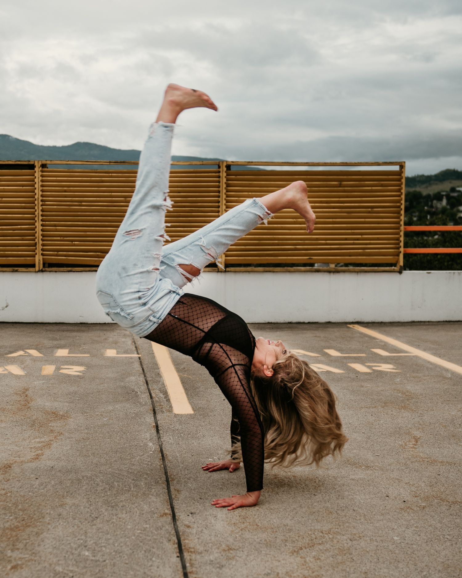 A dancer performs a back-bend handstand on a rooftop with mountains visible in the background under cloudy skies.