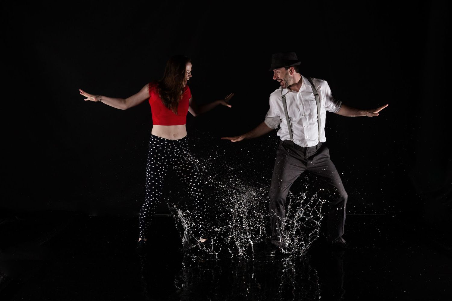 Two dancers perform an energetic duet with water splashing around their feet against a black background.