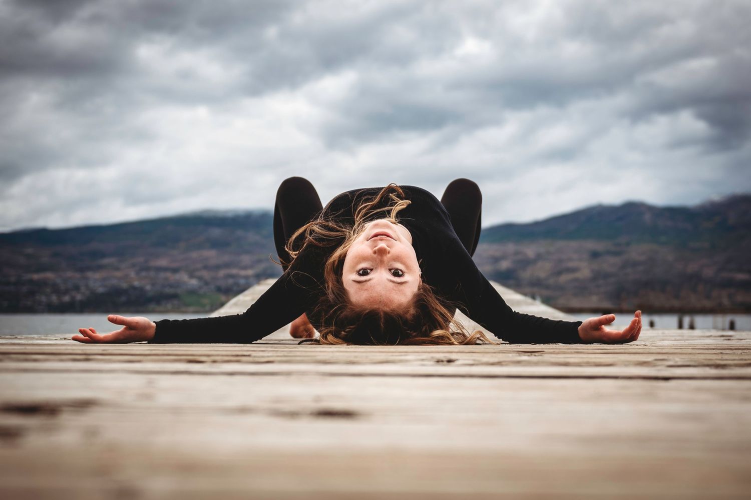 Someone in casual wear lies upside down on a wooden pier with mountains and cloudy skies in the background.