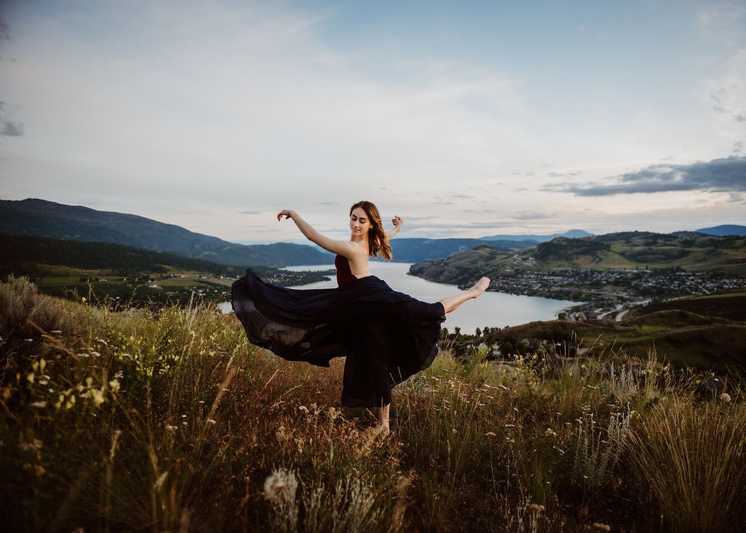 A dancer in a flowing black dress spins gracefully on a hilltop overlooking a scenic lake and mountains at dusk.